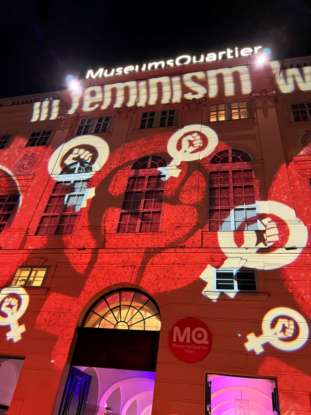 MuseumsQuartier Wien building at night. As part of an exhibit or event, the building has red light projected on it, with the sign for female with a fist raised in the middle of the circle projected in white.